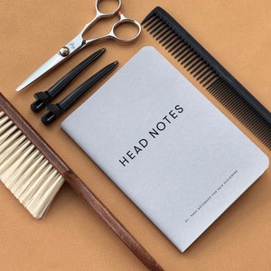 May include: A gray notebook with the text "HEAD NOTES" and the words "84 PAGE NOTEBOOK FOR HAIR DESIGNERS" on the cover. The notebook is surrounded by hairdressing tools, including a pair of scissors, a comb, hair clips, and a brush.