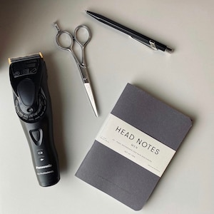 Può includere: Un tagliacapelli elettrico nero con un bordo dorato, un paio di forbici d'argento, una penna nera e un taccuino grigio con il testo "HEAD NOTES MEN PAGE NOTEBOOKS FOR HAIR DESIGNERS SET OF TWO" sulla copertina.