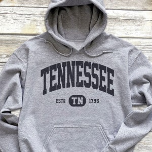 May include: A heather gray hoodie with the word "TENNESSEE" in a black, arched font. Below the word is "ESTD TN 1796". The hoodie has a drawstring hood and a front pocket. The hoodie is on a wooden surface.
