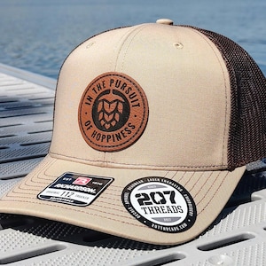 May include: A tan and brown trucker hat with a brown leather patch that says "In the Pursuit of Happiness" with a hop graphic. The hat has a black mesh back and a brown adjustable strap with the words "Richardson 112 Trucker Adjustable" and "207 Threads" printed on it.