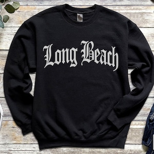 May include: Black crewneck sweatshirt with the words "Long Beach" in a gothic font. The sweatshirt is made of a soft material and is a casual style. The design is printed in white. The sweatshirt is a unisex style.