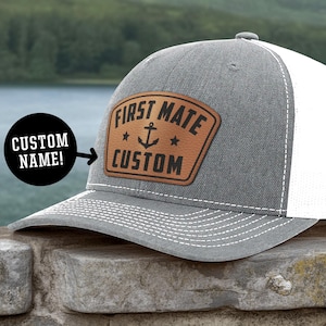 May include: A gray and white trucker hat with a brown leather patch. The patch features the words "FIRST MATE CUSTOM" with an anchor and stars. The hat has a curved brim and a mesh back.