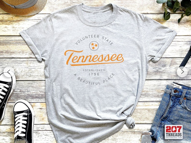 May include: Gray t-shirt with a white graphic print that says "Volunteer State Tennessee Established 1796 A Beautiful Place" with a star in the center of the graphic.