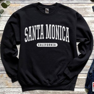 以下が含まれることがあります： 白い文字で「サンタモニカカリフォルニア」と書かれた黒いスウェットシャツ。