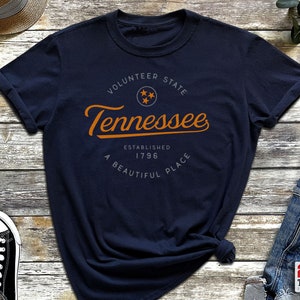 May include: A navy blue t-shirt with a vintage-style graphic design featuring the text "Volunteer State Tennessee Established 1796 A Beautiful Place" in orange with a gray star graphic.