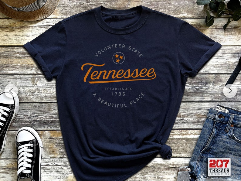 May include: A navy blue t-shirt with a vintage-style graphic design featuring the text "Volunteer State Tennessee Established 1796 A Beautiful Place" in orange with a gray star graphic.