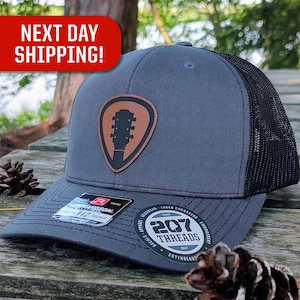 May include: A gray and black trucker hat with a brown leather guitar pick patch. The hat has a black mesh back and a gray front with a black embroidered logo that says "207 Threads".