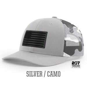 May include: A silver and camo trucker hat with a black American flag patch. The hat has a mesh back and the text "207 Threads" is printed on the back.