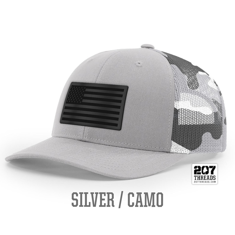 May include: A silver and camo trucker hat with a black American flag patch. The hat has a mesh back and the text "207 Threads" is printed on the back.