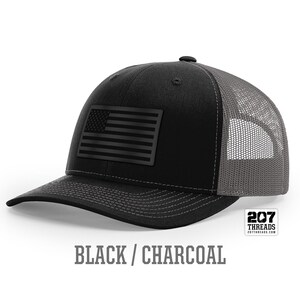 May include: A black and charcoal trucker hat with a black American flag patch on the front. The hat has a mesh back and the text "207 Threads" is printed on the back.