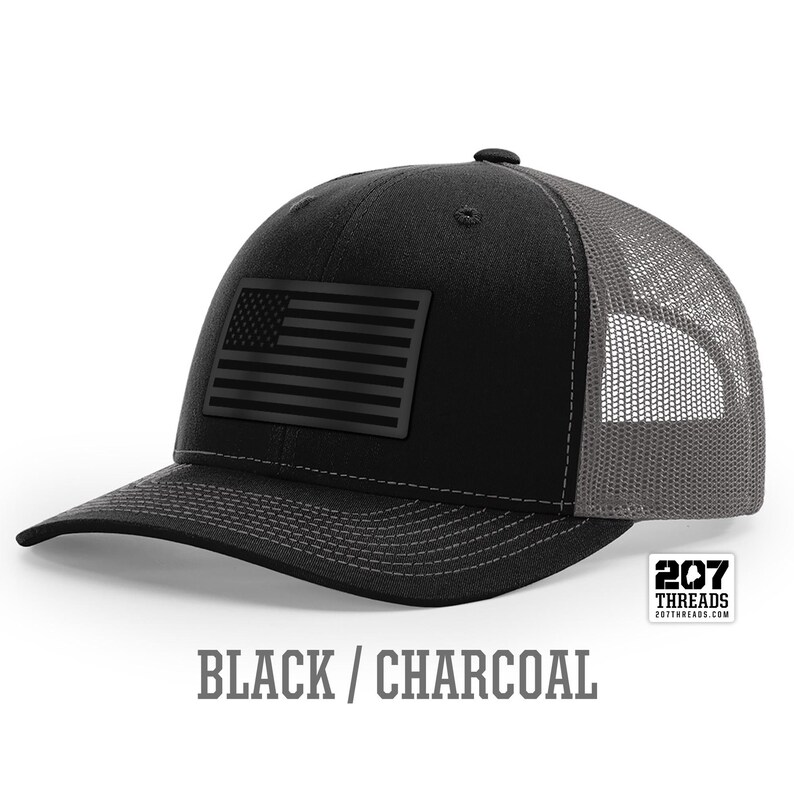 May include: A black and charcoal trucker hat with a black American flag patch on the front. The hat has a mesh back and the text "207 Threads" is printed on the back.