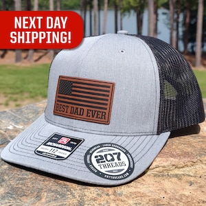 May include: A gray and black trucker hat with a brown leather patch that says "Best Dad Ever" and a small American flag. The hat has a black mesh back and a gray front.