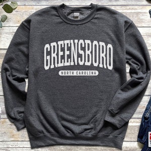 May include: A dark gray sweatshirt with the word "GREENSBORO" in large white block letters across the chest, with "NORTH CAROLINA" printed below. The sweatshirt is a crew neck style.