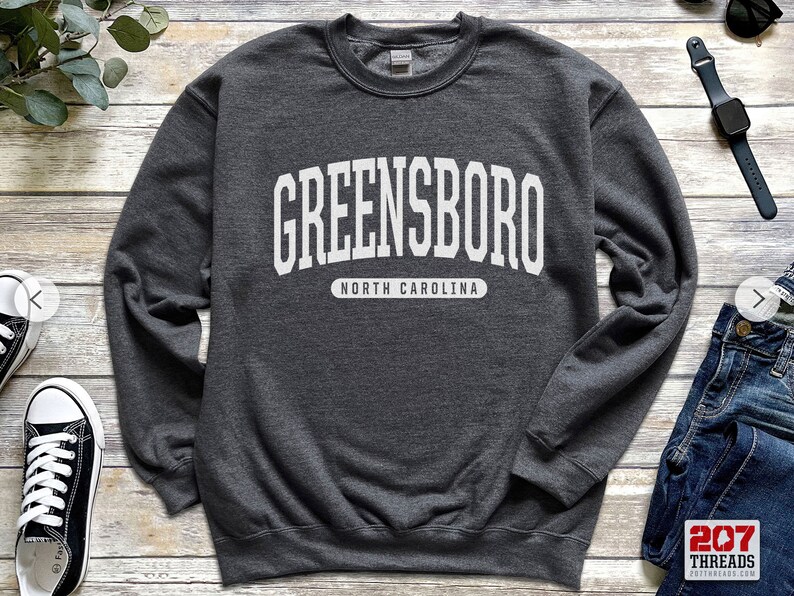 May include: A dark gray sweatshirt with the word "GREENSBORO" in large white block letters across the chest, with "NORTH CAROLINA" printed below. The sweatshirt is a crew neck style.