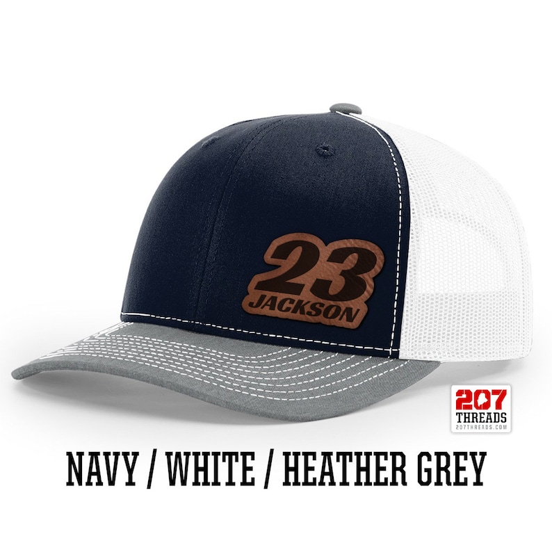 May include: A navy blue, white, and heather grey trucker hat with a brown leather patch that says "23 JACKSON".