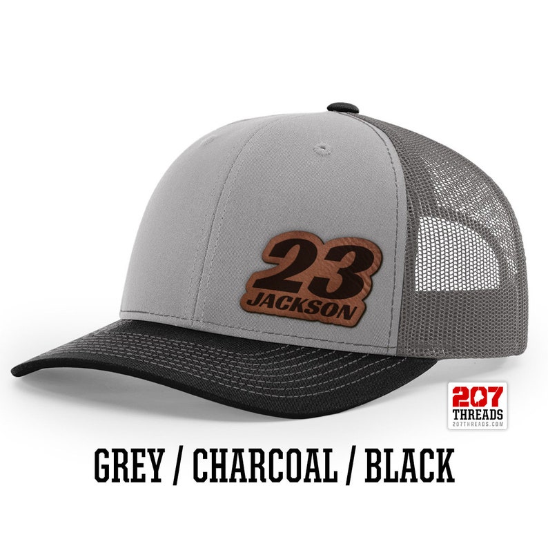 May include: A grey and charcoal trucker hat with a black brim. The hat has a brown leather patch with the number 23 and the name Jackson on it.