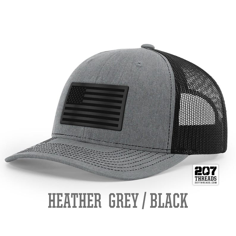 May include: Heather grey trucker hat with a black American flag patch and black mesh back. 207 Threads.