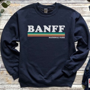 May include: A navy blue sweatshirt with a white graphic that says "BANFF NATIONAL PARK" in a retro style with colorful stripes.