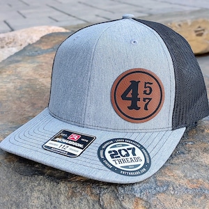 May include: A gray and black trucker hat with a brown leather patch that says "457" in a vintage font. The hat is adjustable and has a black mesh back.