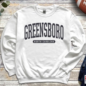 May include: White sweatshirt with the word "GREENSBORO" in dark gray block letters arched across the chest, with "NORTH CAROLINA" below. The sweatshirt is a crew neck style.