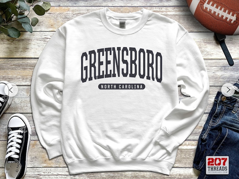 May include: White sweatshirt with the word "GREENSBORO" in dark gray block letters arched across the chest, with "NORTH CAROLINA" below. The sweatshirt is a crew neck style.
