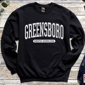 May include: Black sweatshirt with the word "GREENSBORO" in large white block letters across the chest. Below the city name, the text "NORTH CAROLINA" is printed in a smaller white font.