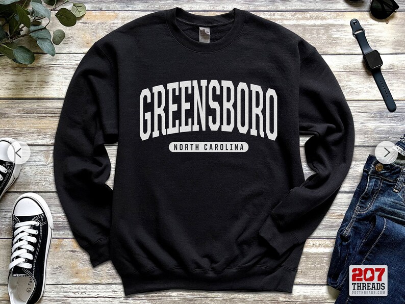 May include: Black sweatshirt with the word "GREENSBORO" in large white block letters across the chest. Below the city name, the text "NORTH CAROLINA" is printed in a smaller white font.