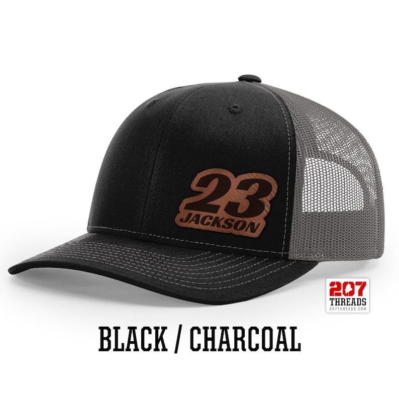 May include: Black and charcoal trucker hat with a brown leather patch that says "23 JACKSON" in white lettering.
