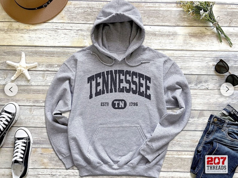 May include: A heather gray hoodie with the word "TENNESSEE" in a black, arched font. Below the word is "ESTD TN 1796". The hoodie has a drawstring hood and a front pocket. The hoodie is on a wooden surface.
