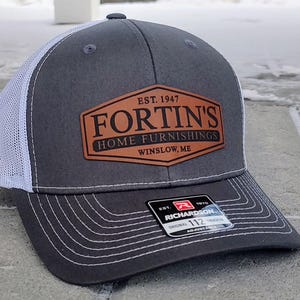 May include: A gray and white trucker hat with a brown leather patch that says "EST. 1947 FORTIN'S HOME FURNISHINGS WINSLOW, ME". The hat is adjustable and has a white mesh back.