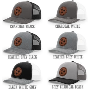 May include: Six trucker hats with a brown leather patch featuring three stars in a circle. The hats are in various color combinations of charcoal, black, white, heather grey, and grey.