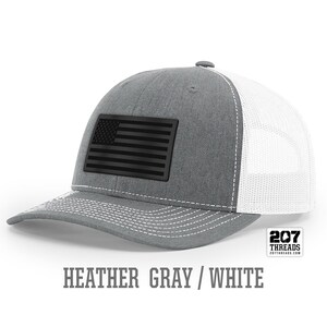 May include: A heather gray and white trucker hat with a black American flag patch. The hat has a mesh back and a curved brim. The text "207 Threads" is on the back of the hat.