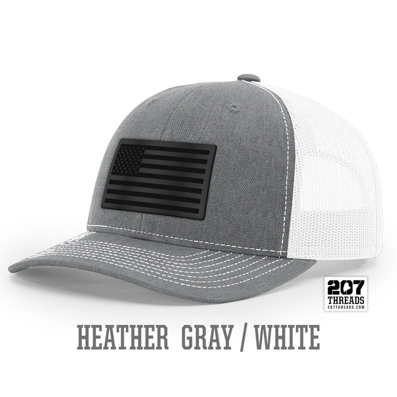 May include: A heather gray and white trucker hat with a black American flag patch. The hat has a mesh back and a curved brim. The text "207 Threads" is on the back of the hat.
