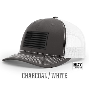 May include: A charcoal gray and white trucker hat with a black American flag patch. The hat has a mesh back and a curved brim. The text "207 Threads" is printed on the back of the hat.