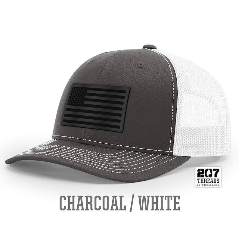 May include: A charcoal gray and white trucker hat with a black American flag patch. The hat has a mesh back and a curved brim. The text "207 Threads" is printed on the back of the hat.
