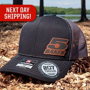 May include: Black and gray trucker hat with a brown leather patch that says "5 BAKER" on it. The hat has a white logo that says "207 THREADS" on the side.