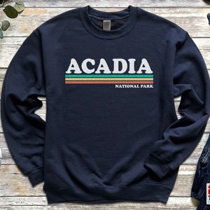 May include: A navy blue sweatshirt with a white graphic that says "Acadia National Park" with a retro-style striped design in green, orange, and yellow.