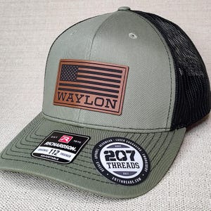 May include: A green and black trucker hat with a brown leather patch that says "WAYLON" and a small American flag. The hat also has a black and white label that says "207 THREADS" and a red and white label that says "RICHARDSON".