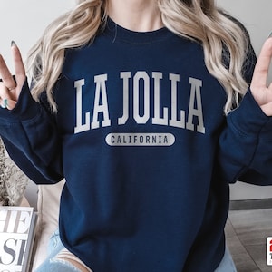 May include: Navy blue sweatshirt with the text "LA JOLLA CALIFORNIA" in white block letters. The sweatshirt is a crew neck style and has a relaxed fit. The text is centered on the chest.