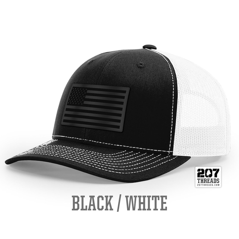 May include: Black and white trucker hat with a black American flag patch on the front. The hat has a white mesh back and a black brim. The text "207 Threads" is printed on a white tag attached to the hat.