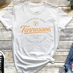 May include: White t-shirt with a graphic design featuring the text "Volunteer State Tennessee Established 1796 A Beautiful Place" in orange and gray text with a star graphic.