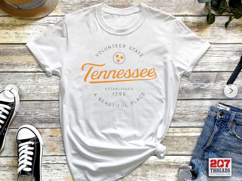 May include: White t-shirt with a graphic design featuring the text "Volunteer State Tennessee Established 1796 A Beautiful Place" in orange and gray text with a star graphic.