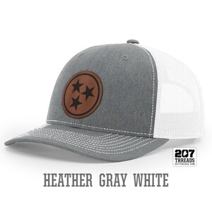 May include: A heather gray and white trucker hat with a brown leather patch featuring three black stars. The patch is a circle with a white outline. The hat has a white mesh back and a gray front. The text "207 Threads" is on a white tag attached to the hat.