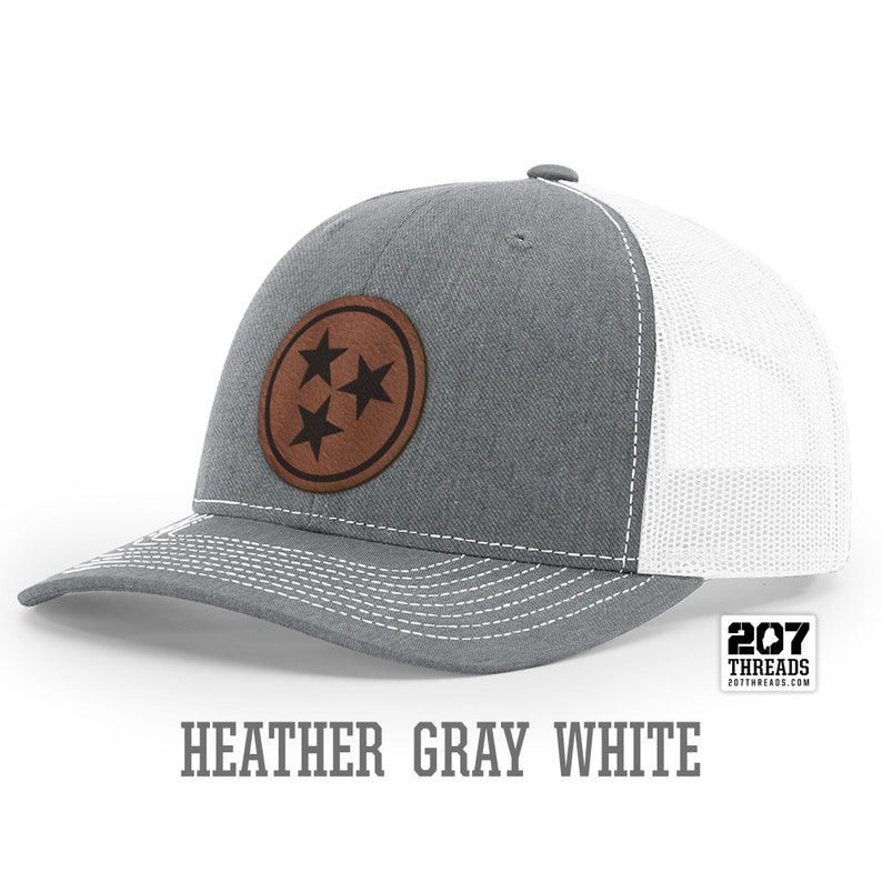 May include: A heather gray and white trucker hat with a brown leather patch featuring three black stars. The patch is a circle with a white outline. The hat has a white mesh back and a gray front. The text "207 Threads" is on a white tag attached to the hat.