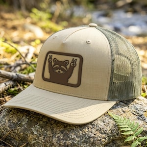 May include: A beige trucker hat featuring a rectangular patch on the front. The patch displays a dark brown graphic of a raccoon making a peace sign with both paws. The hat has a solid beige front panel and a dark olive green mesh back panel.