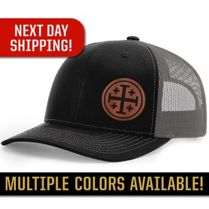May include: Black and gray trucker hat with a brown leather patch featuring a cross with four smaller crosses. The patch is centered on the front of the hat.