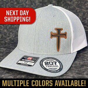 May include: A gray and white trucker hat with a brown leather cross design. The hat has a mesh back and is adjustable. The hat has the text "207 Threads" on the side.