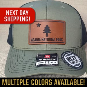 May include: Olive green and black trucker hat with a leather patch featuring a star and tree graphic, and the text "ACADIA NATIONAL PARK." The hat has the text "RICHARDSON ORIGINAL 112 TRUCKER ADJUSTABLE" on the front.