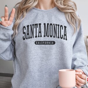 以下が含まれることがあります： 前面に「SANTA MONICA CALIFORNIA」とプリントされたグレーのスウェットシャツです。