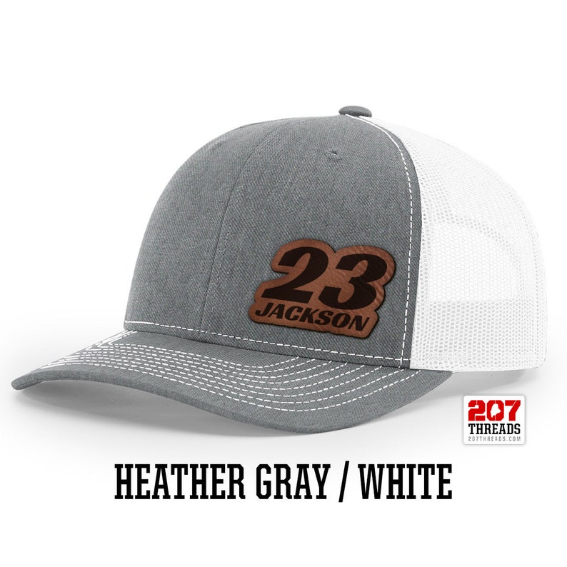 May include: A heather gray and white trucker hat with a brown leather patch that says "23 JACKSON" on it.